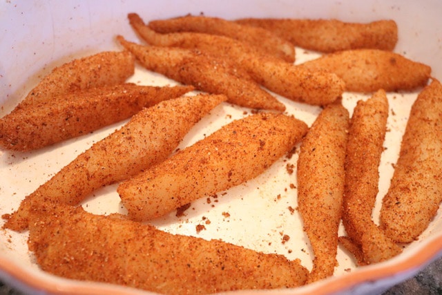 Strips of raw tilapia coated in a spice mixture in a white casserole dish.