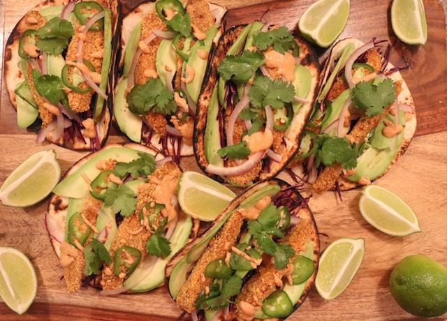 Six fish tacos on a wooden serving board next to lime wedges and a whole lime.