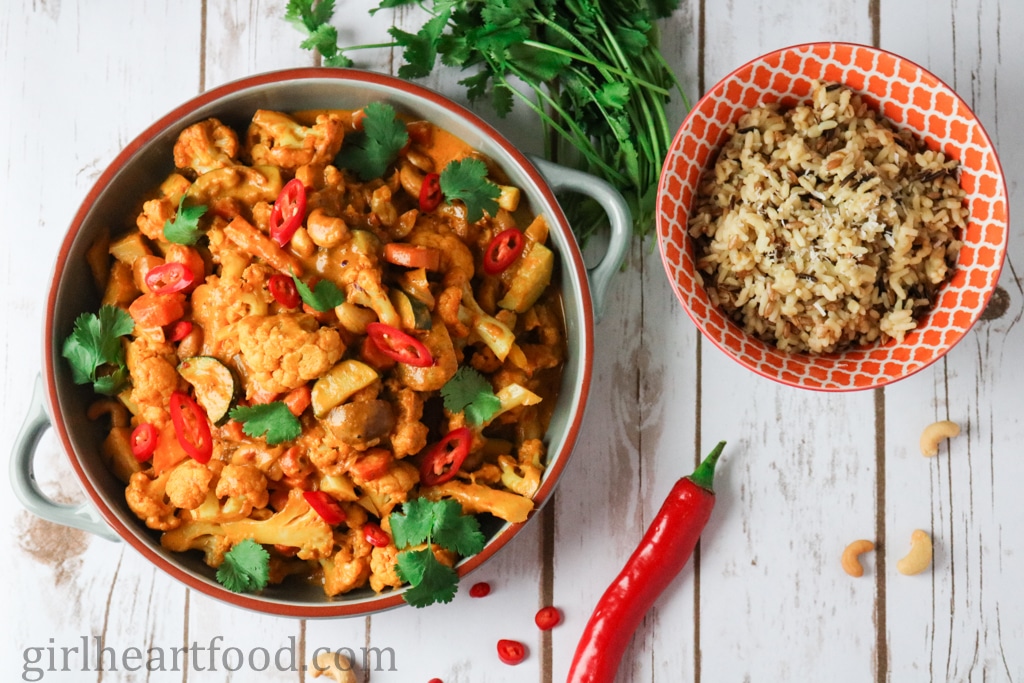 Overhead shot of veggie korma in a dish alongside a bowl of rice, cilantro and chili pepper.