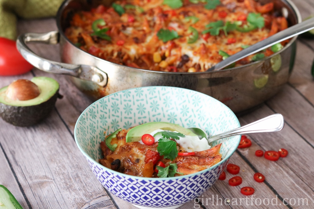 A portion of chili pasta bake in a bowl with the pan of pasta bake behind it.