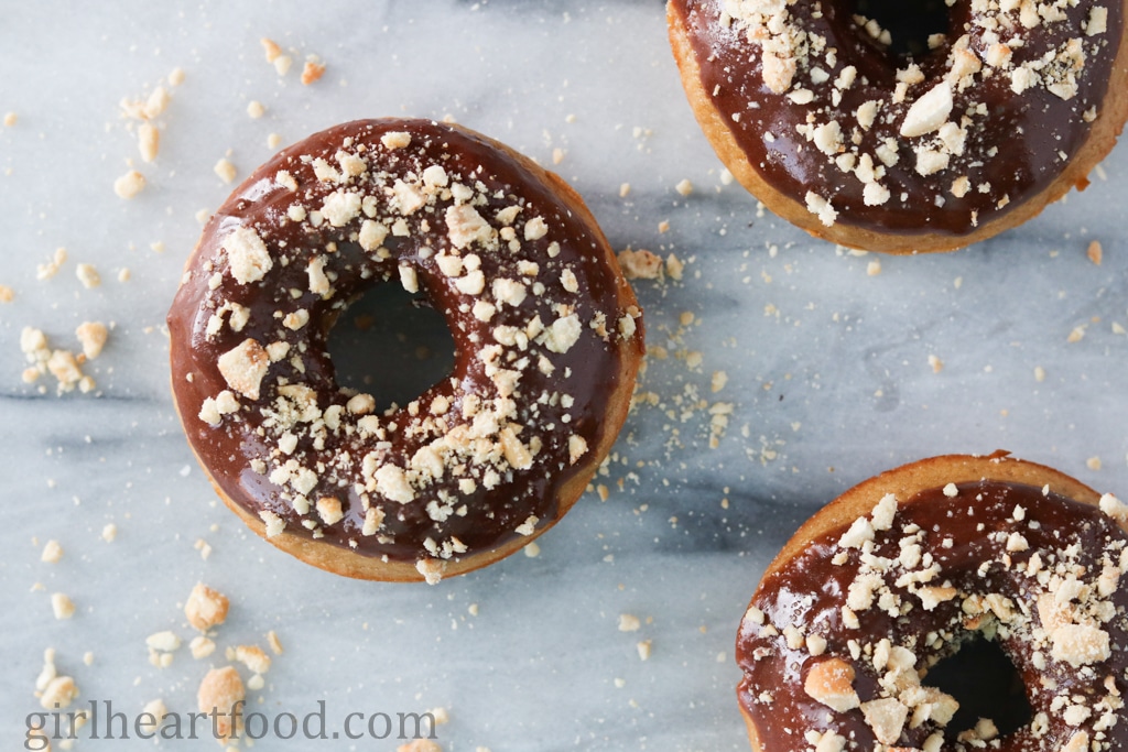 Three baked donuts with glaze and crushed nuts on a marble board.