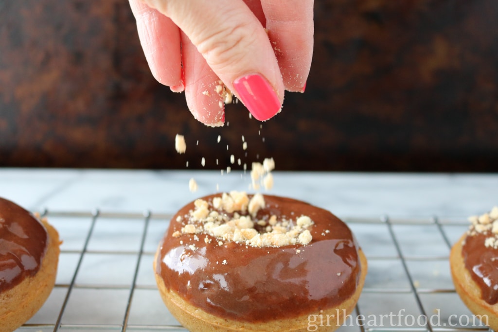 A hand sprinkling crushed peanuts onto glazed donuts.