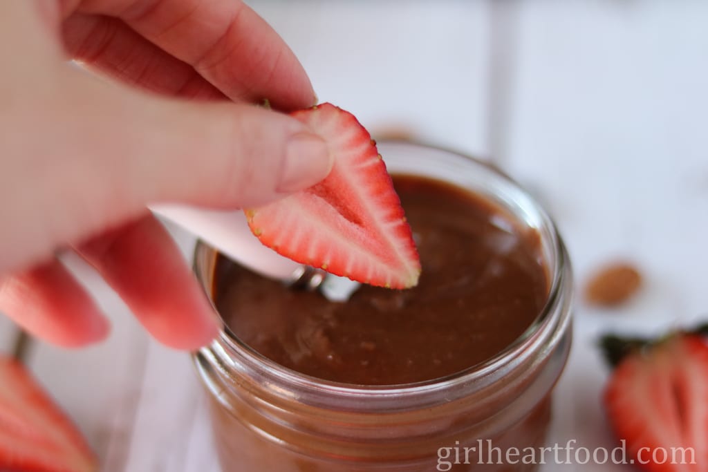 Hand dipping a strawberry into a jar of chocolate almond butter.