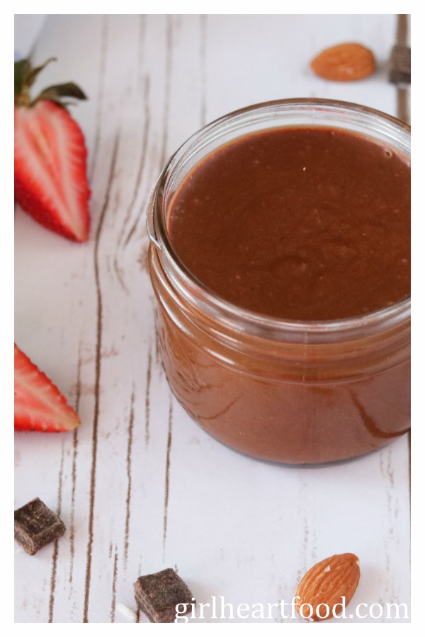 Jar of chocolate almond butter next to a cut strawberry, chocolate chunks and an almond.