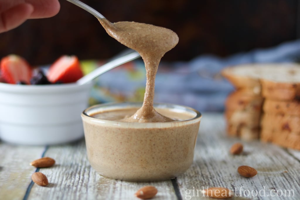 Hand holding a spoonful of roasted almond butter, dripping into a jar underneath it.