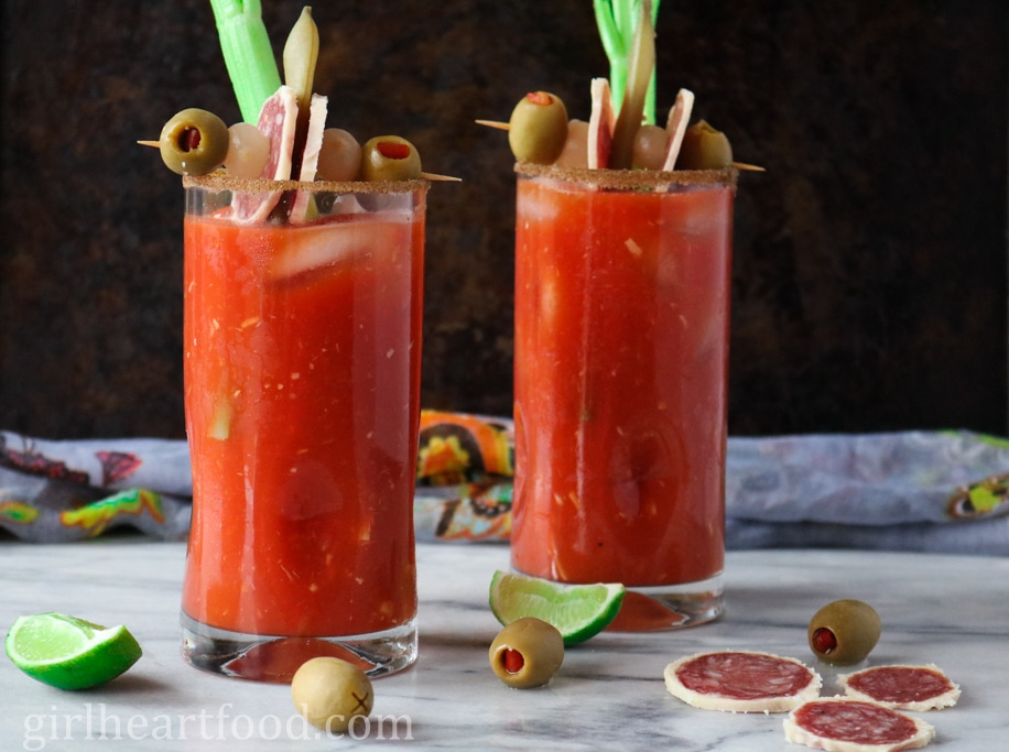 Two glasses of Caesar cocktail, each garnished with a snack and celery stick.
