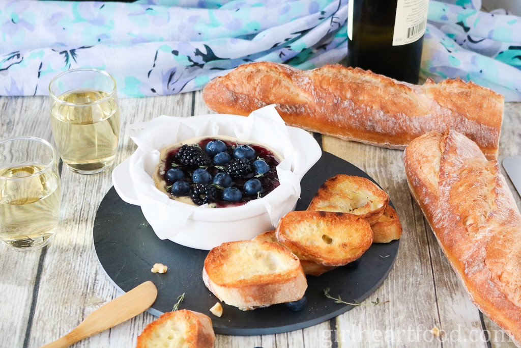 Dish of baked camembert topped with berries, and surrounded by bread and glasses of wine.