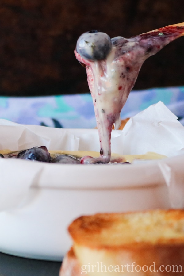 Spreader lifting up cheesy camembert with blueberry from a dish.