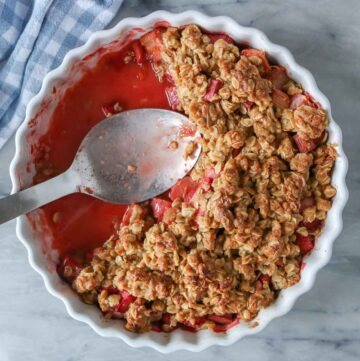 Partially eaten strawberry rhubarb crisp in a serving dish with a serving spoon resting in it.