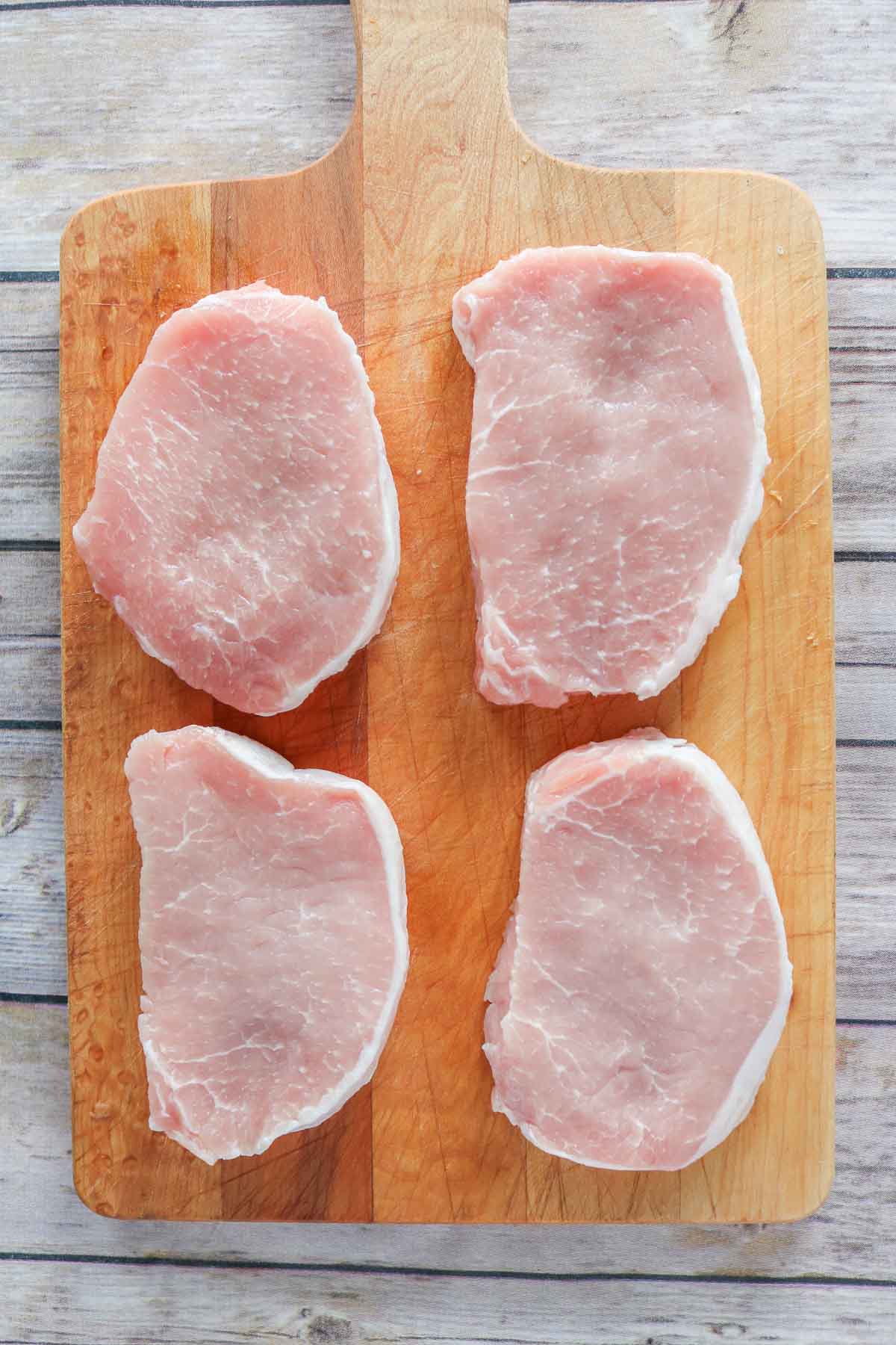 Four uncooked boneless pork loin chops on a wooden board.