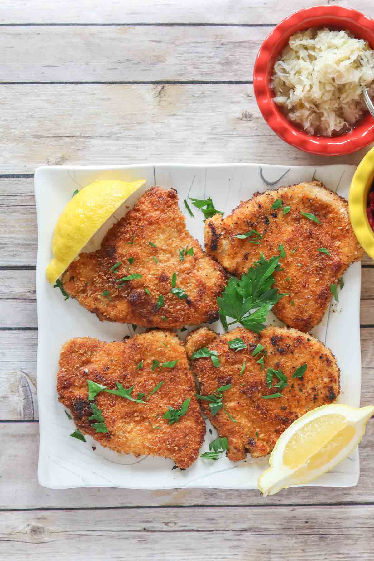Four portions of pork schnitzel on a plate with parsley and lemon next to a dish of sauerkraut.