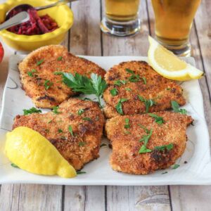Four portions of pork schnitzel on a plate with parsley and lemon wedges.