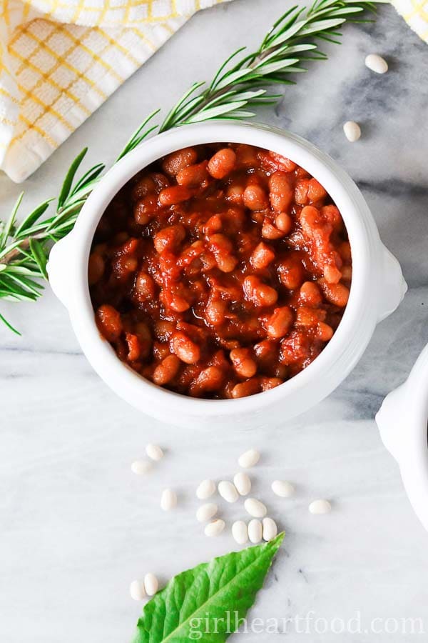 Bowl of homemade baked beans next to fresh rosemary, a bay leaf and dried beans.