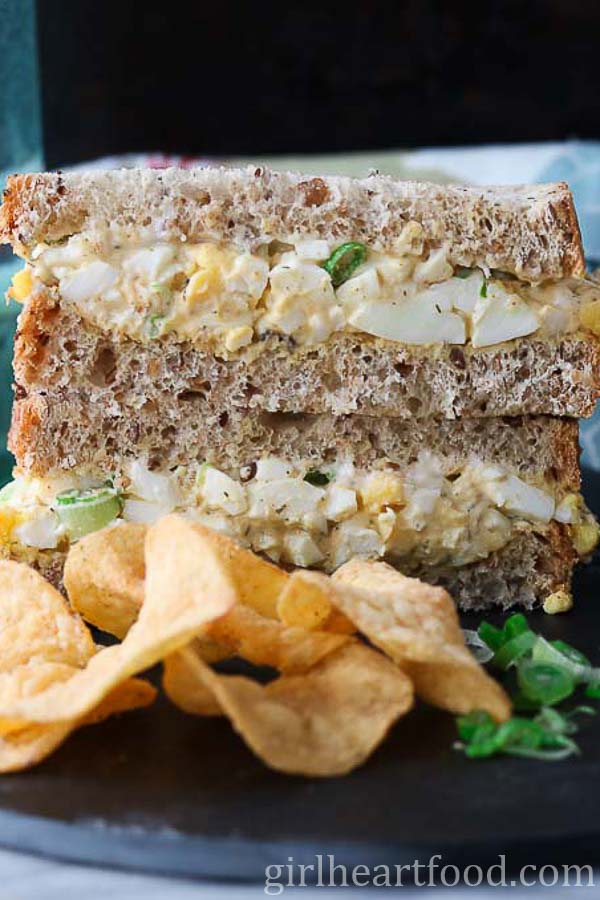 Stack of two halves of an egg salad sandwich next to potato chips.