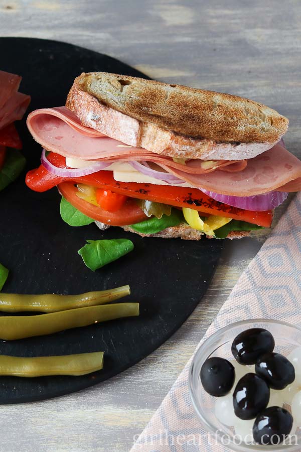 A mortadella sandwich next to pickled green beans, black olives and pickled onions.