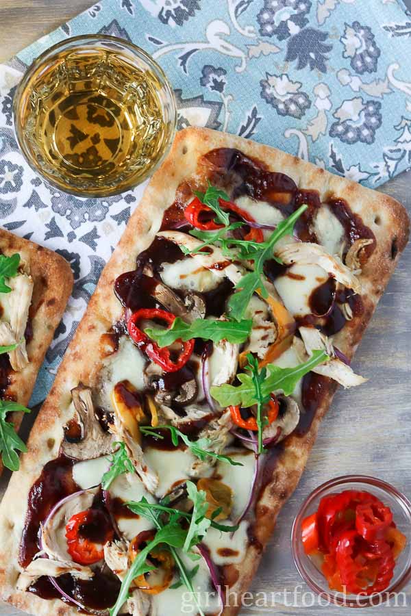 BBQ chicken flatbread pizza next to a glass of wine and dish of hot peppers.