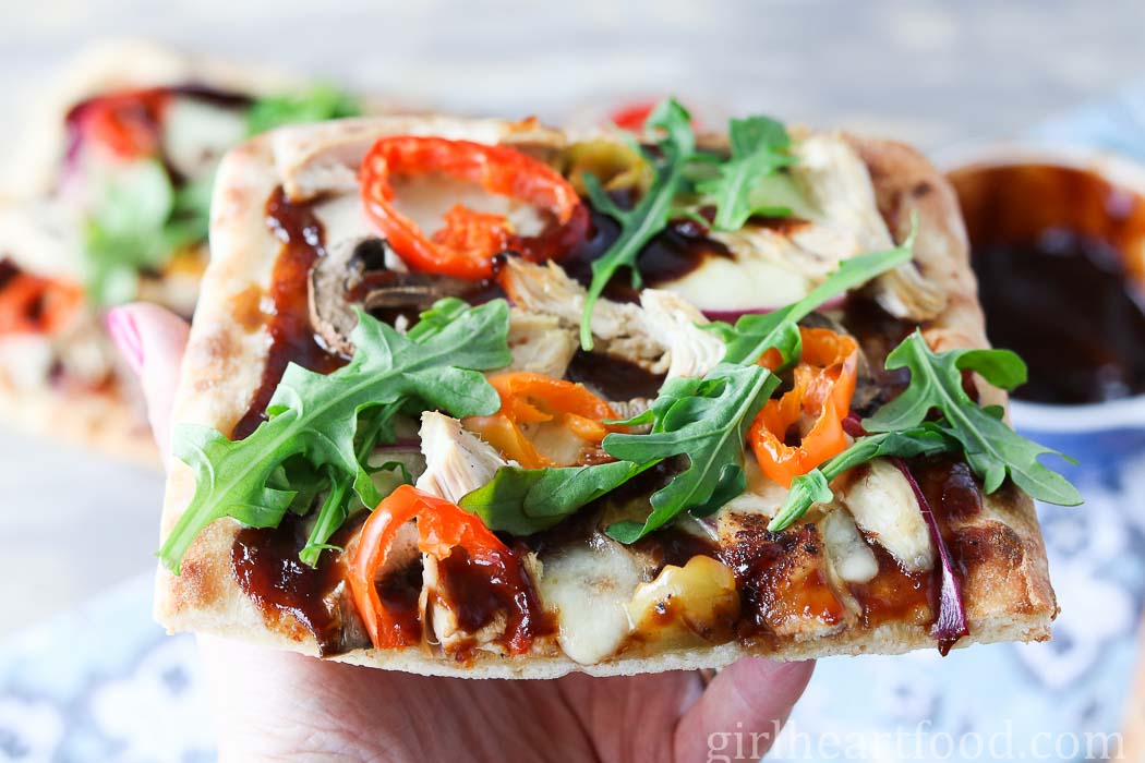 Hand holding a piece of BBQ chicken flatbread pizza.