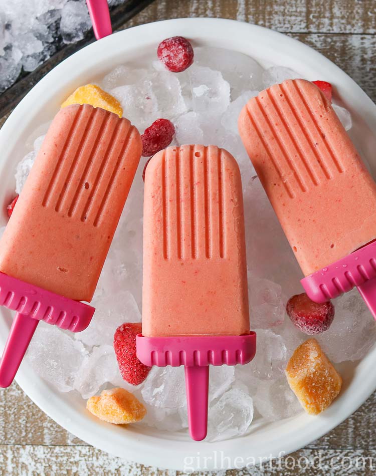 Three strawberry mango ice pops on top of a bowl of ice cubes.