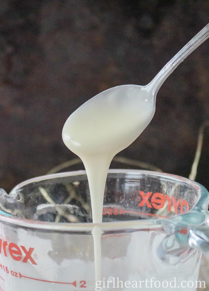 A spoonful of icing sugar glaze above a measuring cup, dripping into it.