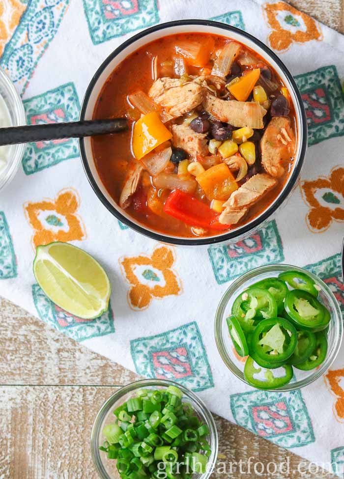 Bowl of chicken taco soup next to dishes of sliced jalapeno and green onion and a wedge of lime.