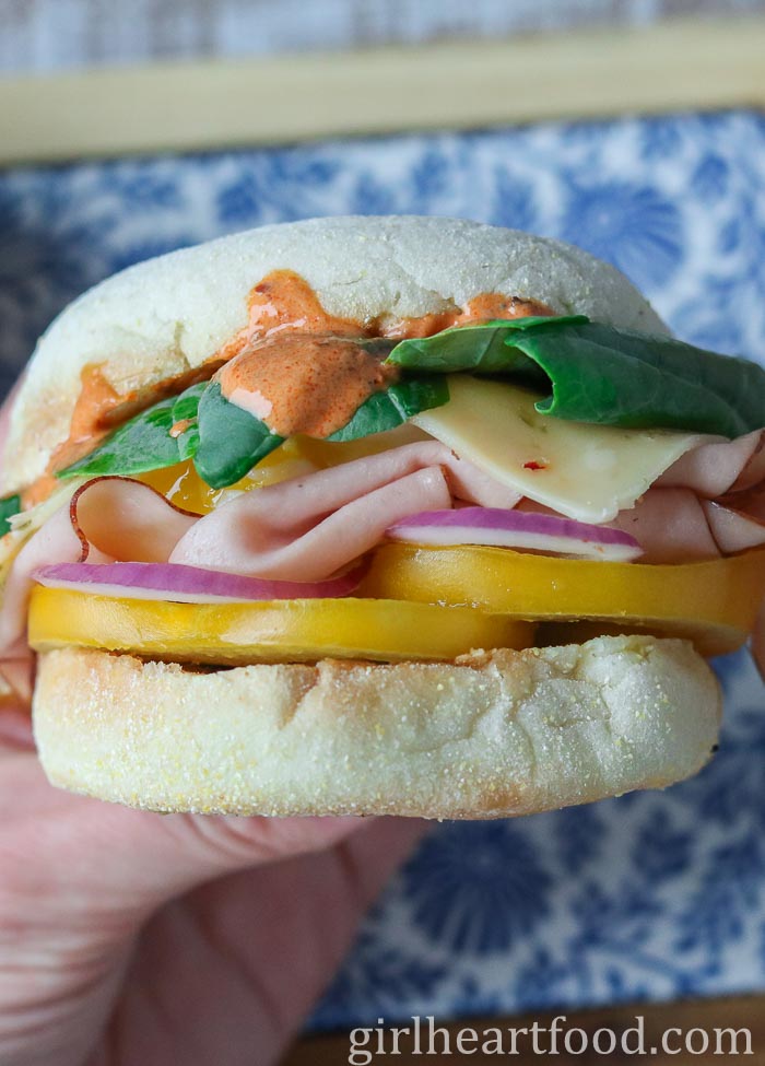 Hand holding a ham and egg sandwich with toppings and sauce.