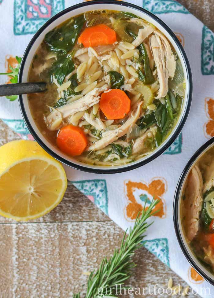 Bowl of lemon chicken orzo soup next to half a lemon and fresh rosemary.