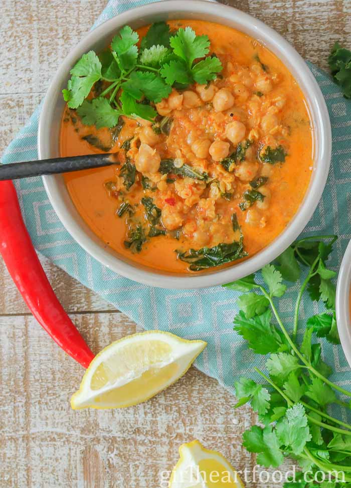 Bowl of red lentil chickpea soup next to a red chili pepper, lemon wedge and cilantro.