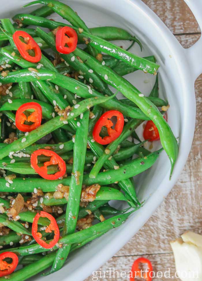 Garlicky green beans in a dish topped with sliced chili pepper and sesame seeds.