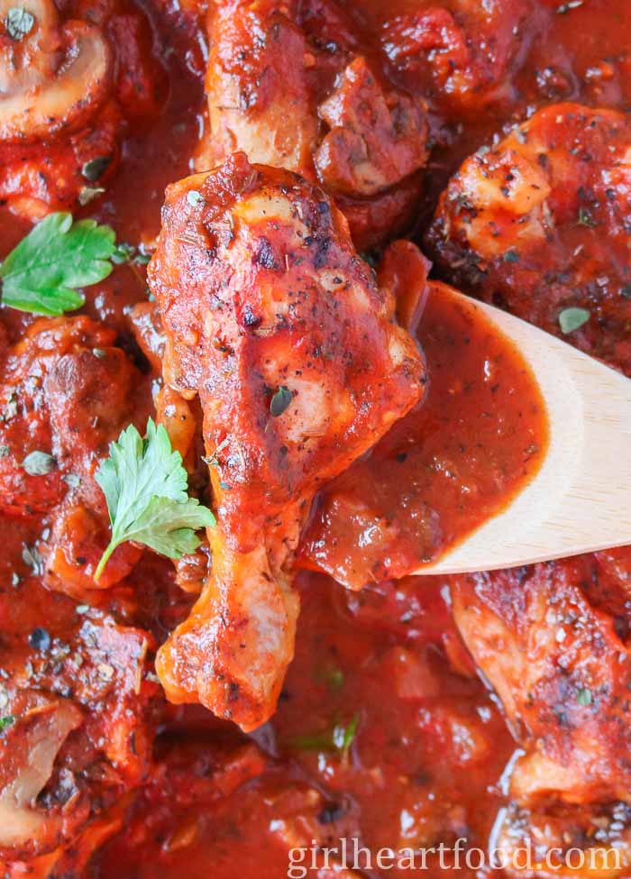 Wooden spoon scooping up a chicken leg with tomato sauce from a pan.