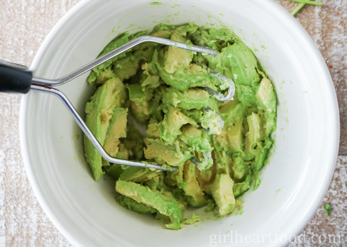 Mashing avocado in a white bowl with a potato masher.