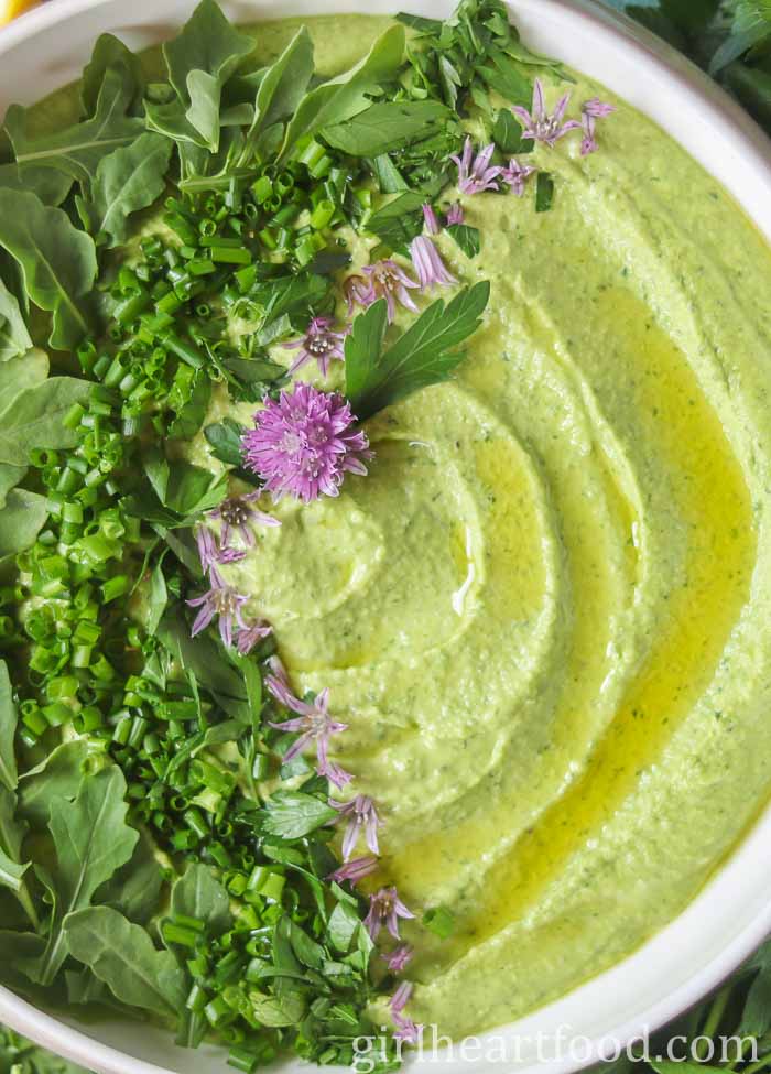 Close-up of a bowl of green goddess hummus garnished with herbs, oil and chive flowers.