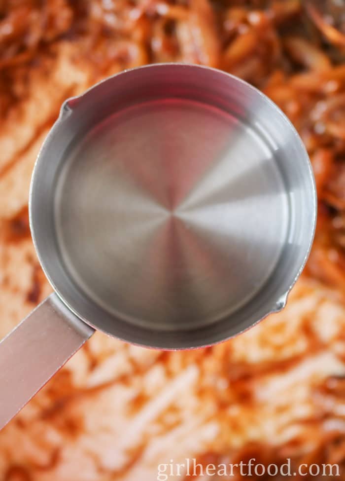 A measuring cup of water about to be poured into a pan of caramelized onions.