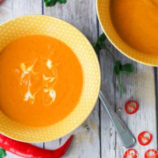 Two bowls of curry carrot soup next to chili peppers, cilantro and a carrot.