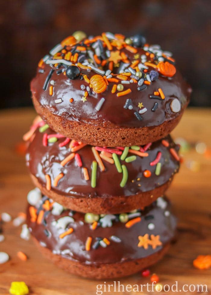 Stack of three pumpkin chocolate donuts with ganache and sprinkles.
