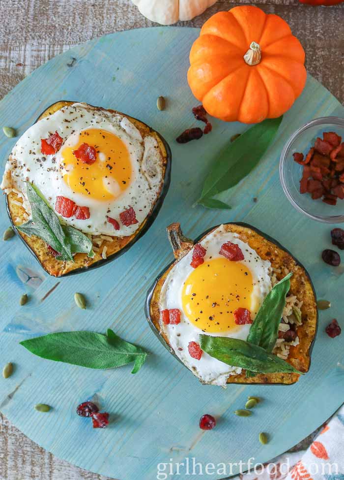Two portions of roasted stuffed acorn squash, each topped with an egg, bacon and sage.
