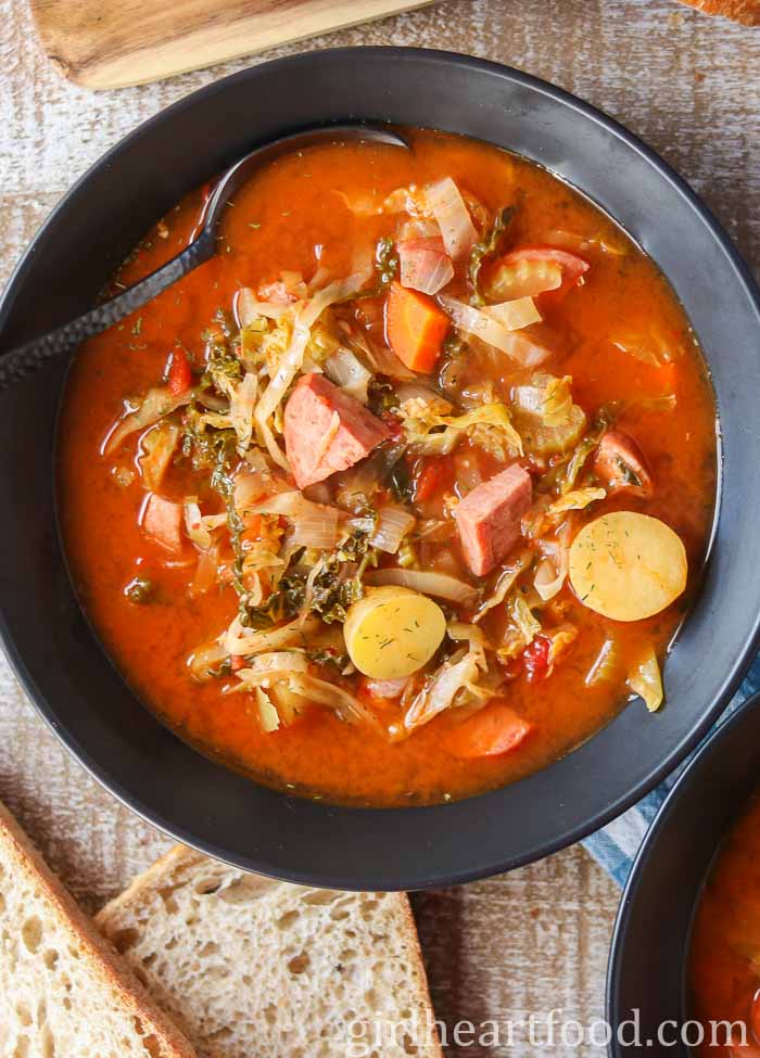 Bowl of cabbage, sausage and potato soup alongside some bread.