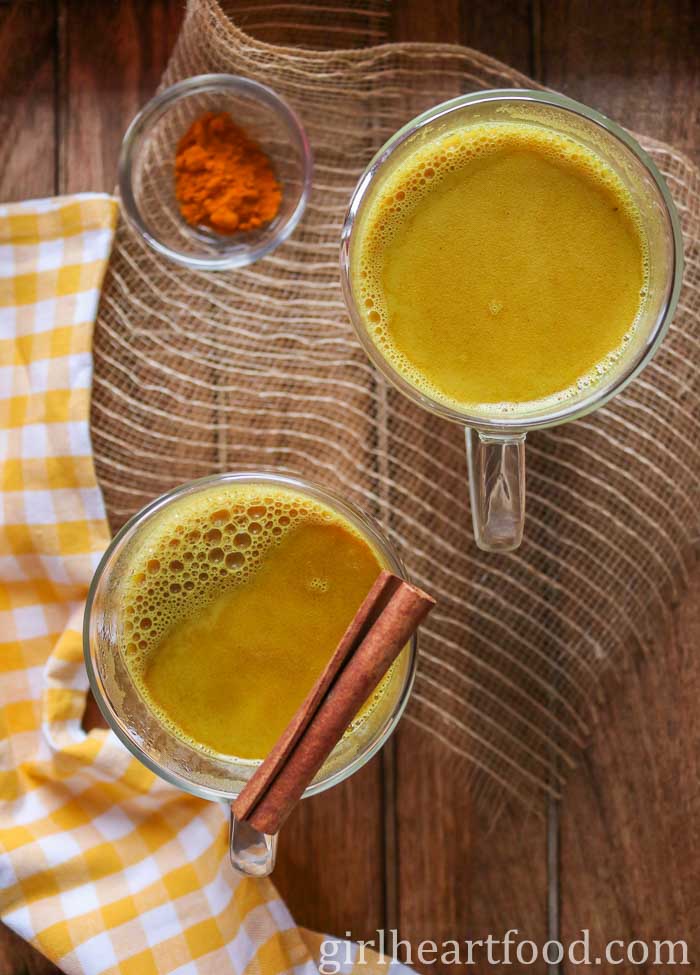 Two mugs of turmeric golden milk next to a small dish of ground turmeric.