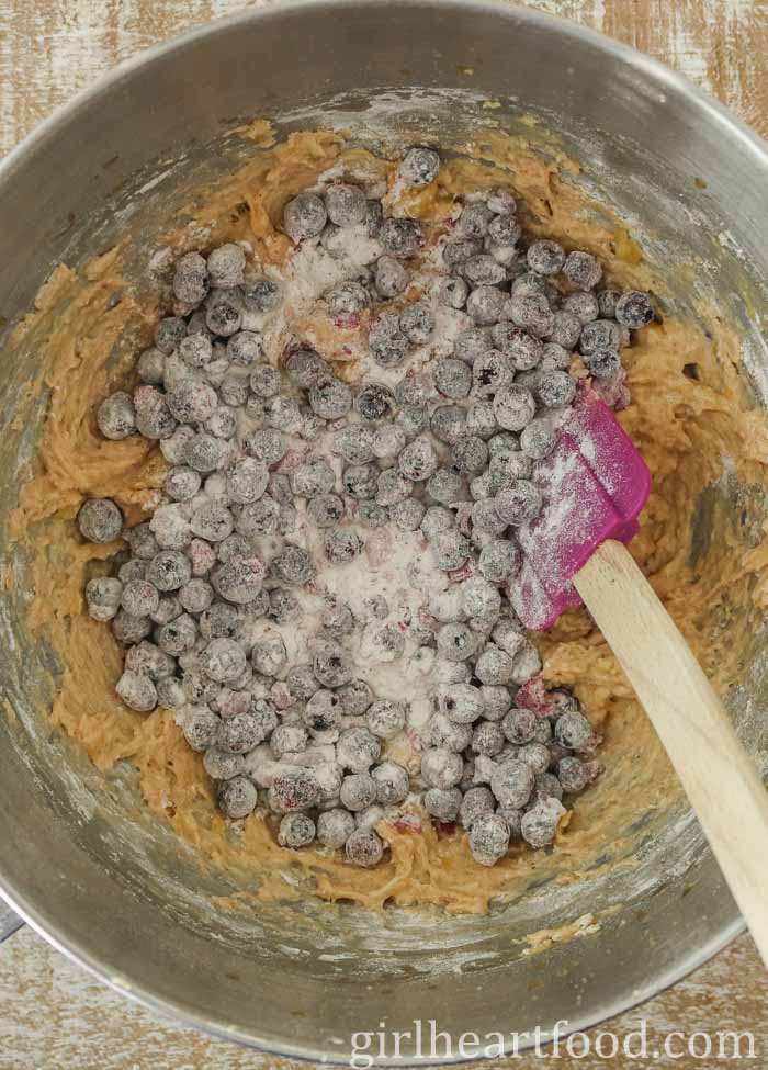 Batter and flour-dusted blueberries in a mixing bowl being stirred by a spatula.