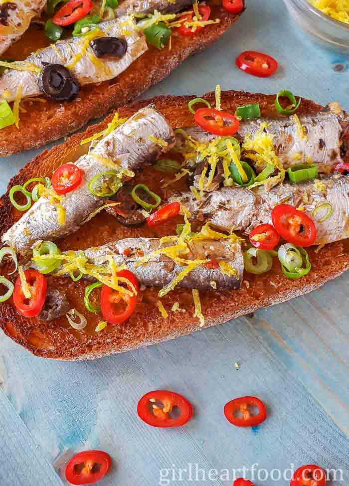 Two slices of toast with sardines, chili pepper, lemon zest and green onion on top.