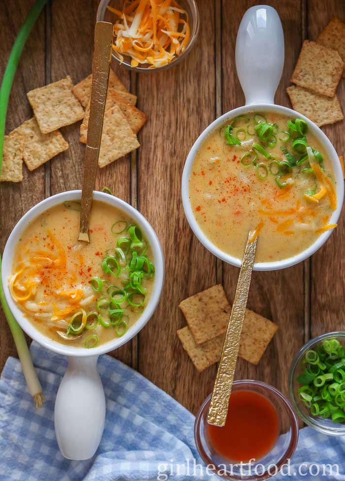 Two bowls of cheese & potato soup next to crackers, green onion, hot sauce & cheese.