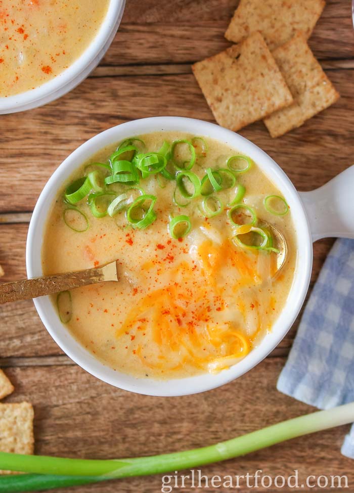 Bowl of cheese and potato soup next to crackers, green onion and a tea towel.