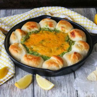 Spinach artichoke dip and biscuits in a skillet next to lemon, a tea towel & Parmesan.