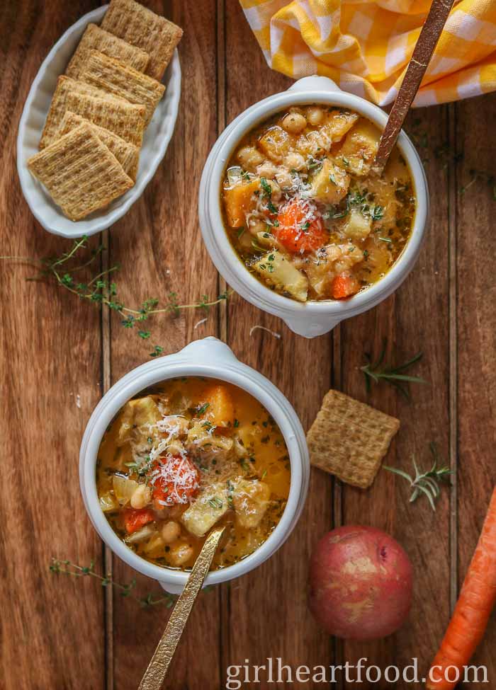 Two bowls of chunky vegetable soup next to crackers and veggies.