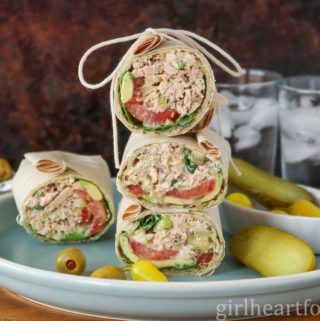 Stack of three tuna salad wraps next to another wrap, pickle, olives and pickled hot pepper.