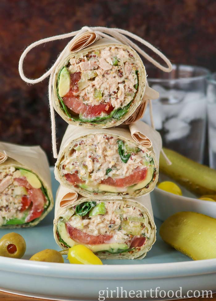 Stack of three tuna salad wraps next to another wrap, pickle, olives and pickled hot pepper.