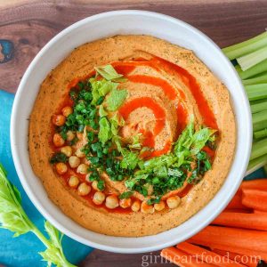 Bowl of buffalo hummus garnished with toppings next to carrot sticks and celery sticks.