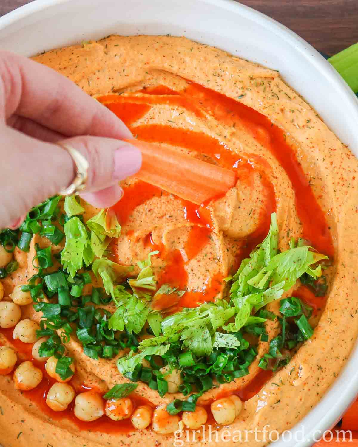 Hand dipping a carrot stick into a bowl of buffalo hummus.