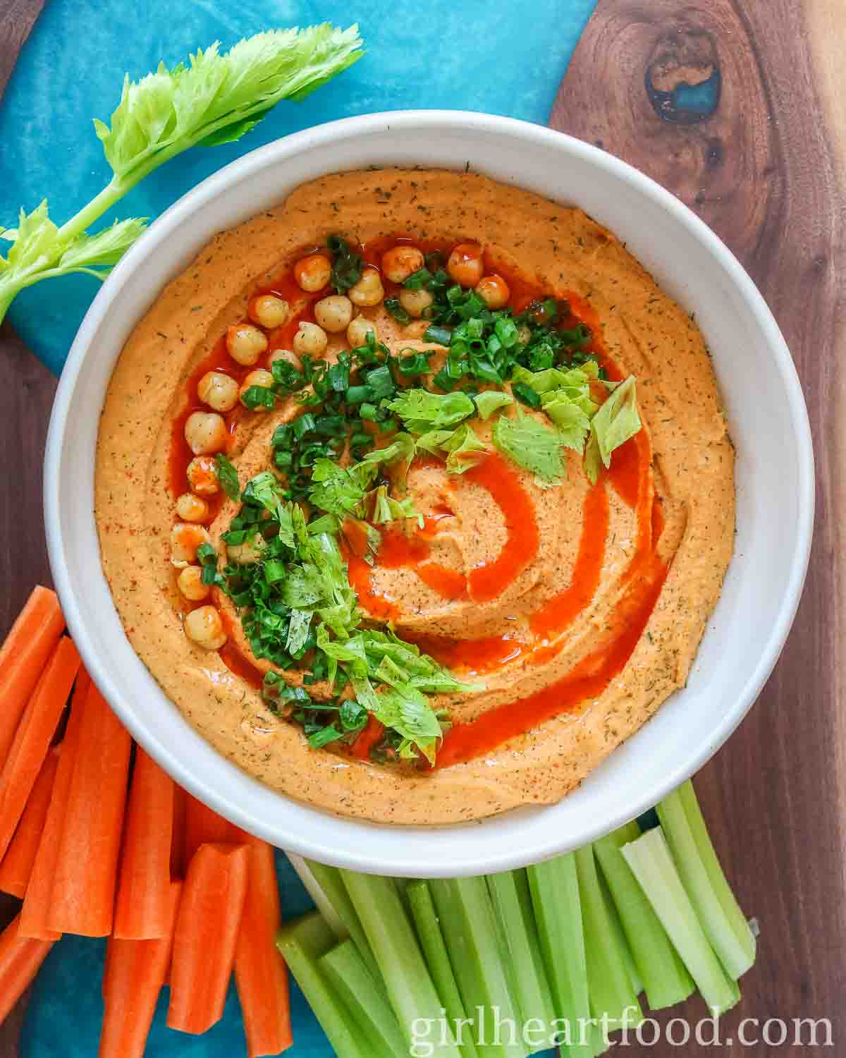 Bowl of buffalo hummus garnished with toppings, next to carrot sticks and celery sticks.