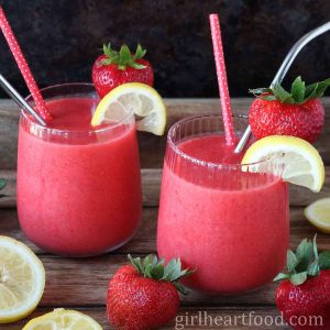 Two glasses of frozen strawberry lemonade alongside strawberries and lemon.