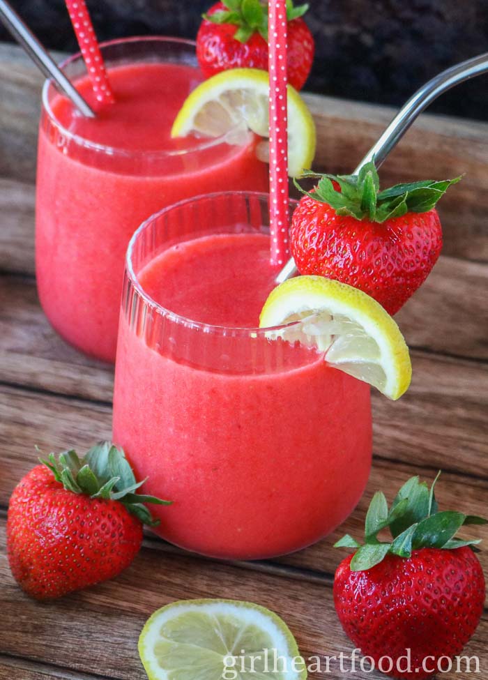 Two glasses of frozen strawberry lemonade, one in front of the other, next to strawberries & lemon.